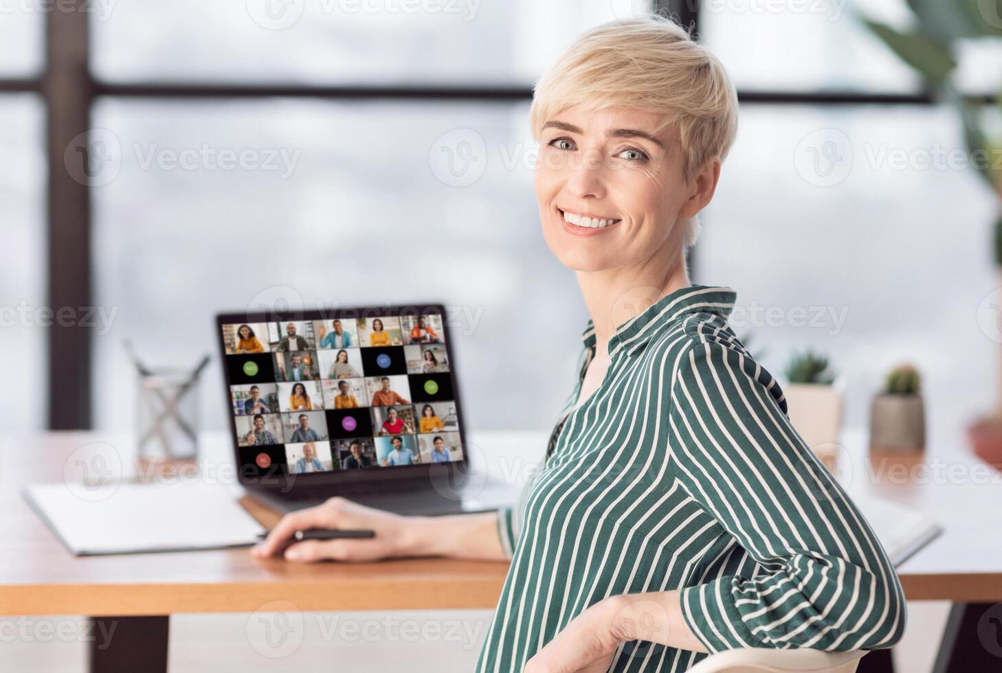 Mature woman with short blonde hair sits at a desk in an office setting with a laptop open in front of her, looking at the camera and smiling, with her right hand resting on the back of her chair photo