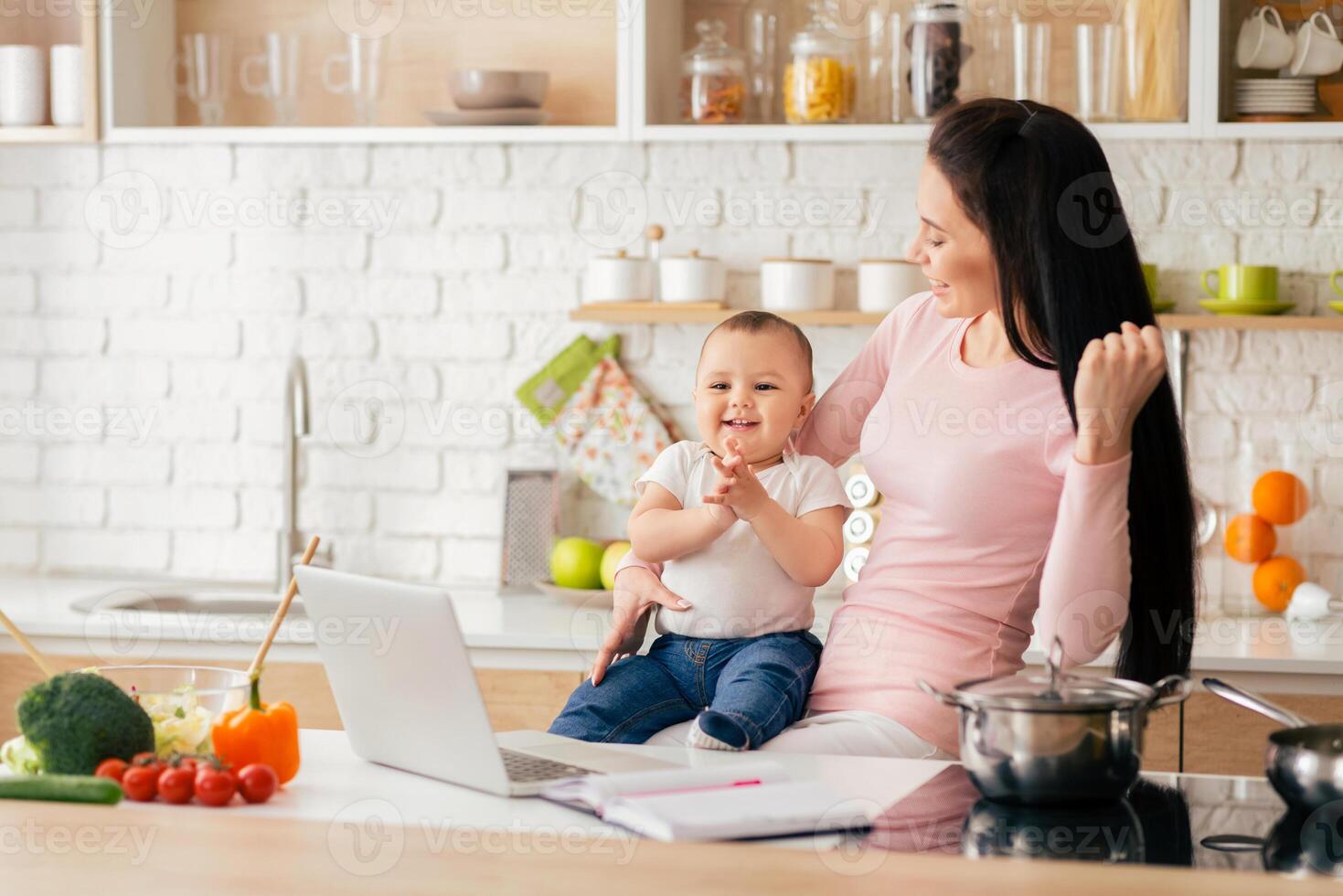 A mother is holding her smiling baby while working at a kitchen counter. The kitchen is modern and well-organized with fresh vegetables and cooking utensils on the counter. photo