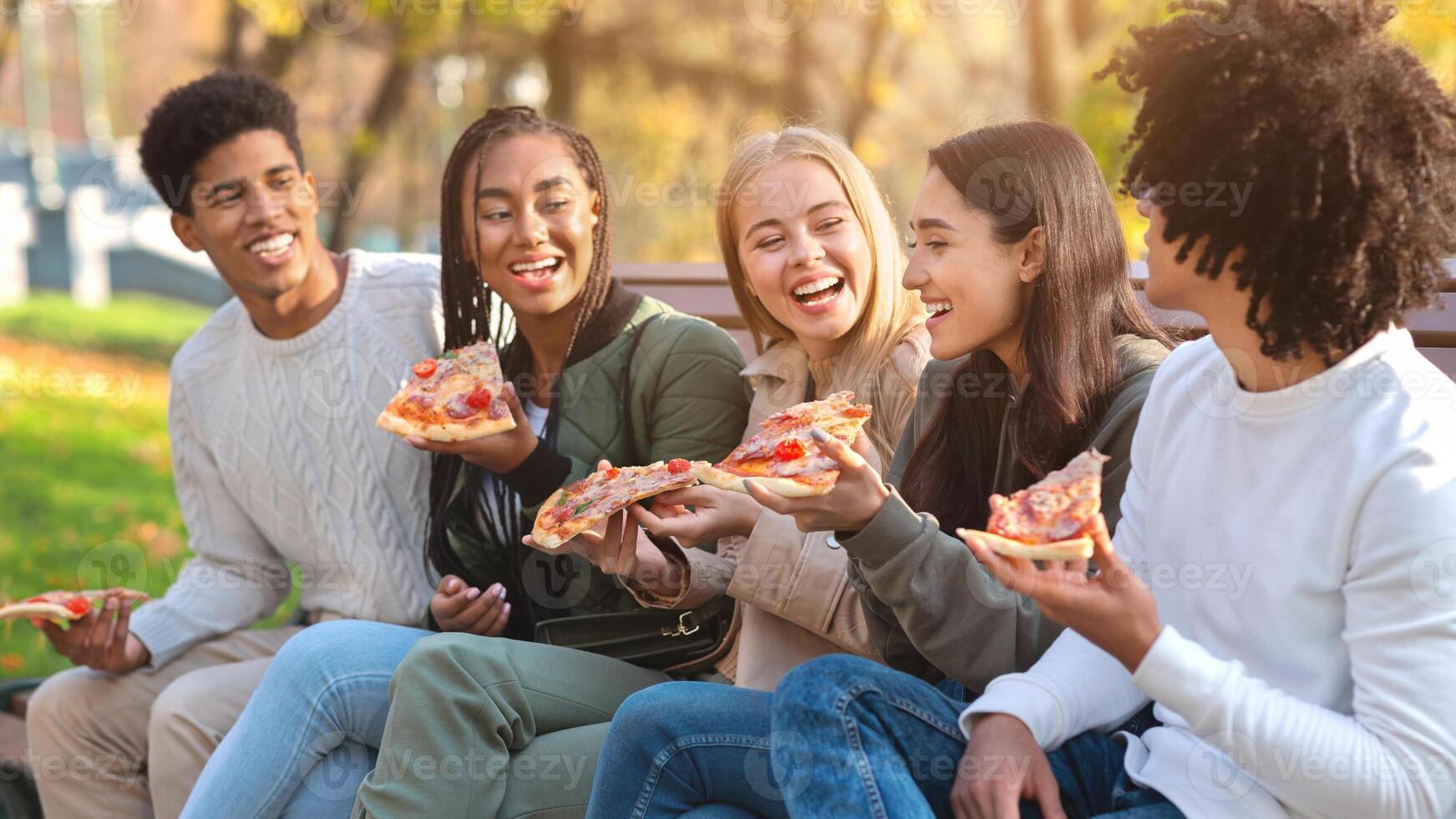 Hungry laughing international teenagers enjoying pizza while spending time in park photo