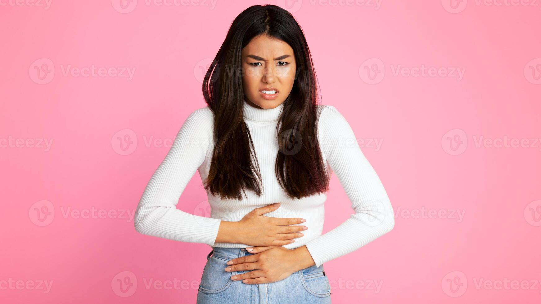 A young Asian woman with long dark hair is standing in front of a pink background. She is holding her stomach with both hands, looking distressed and pained. photo