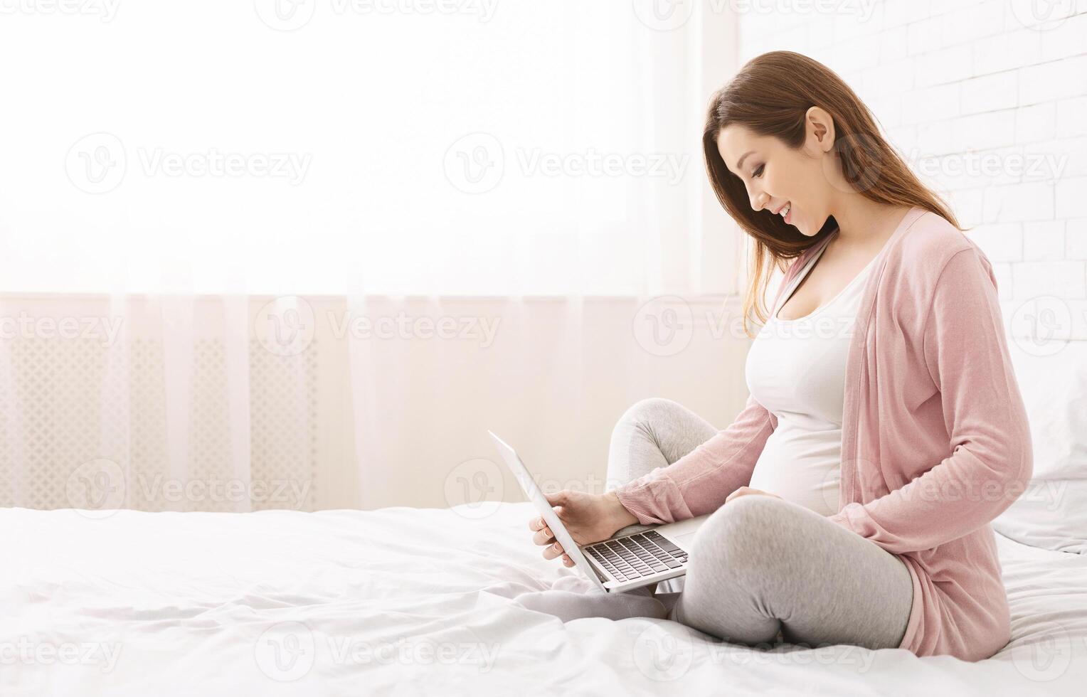 A woman is sitting on a bed indoors, focused on using a laptop computer. She appears to be engaged in online activities, copy space photo