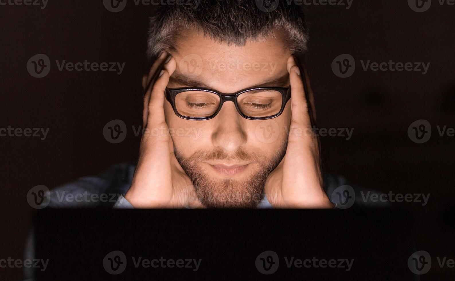 A man wearing glasses concentrates intensely while working on his laptop late at night. His hands are on his head, indicating deep thought or stress. photo