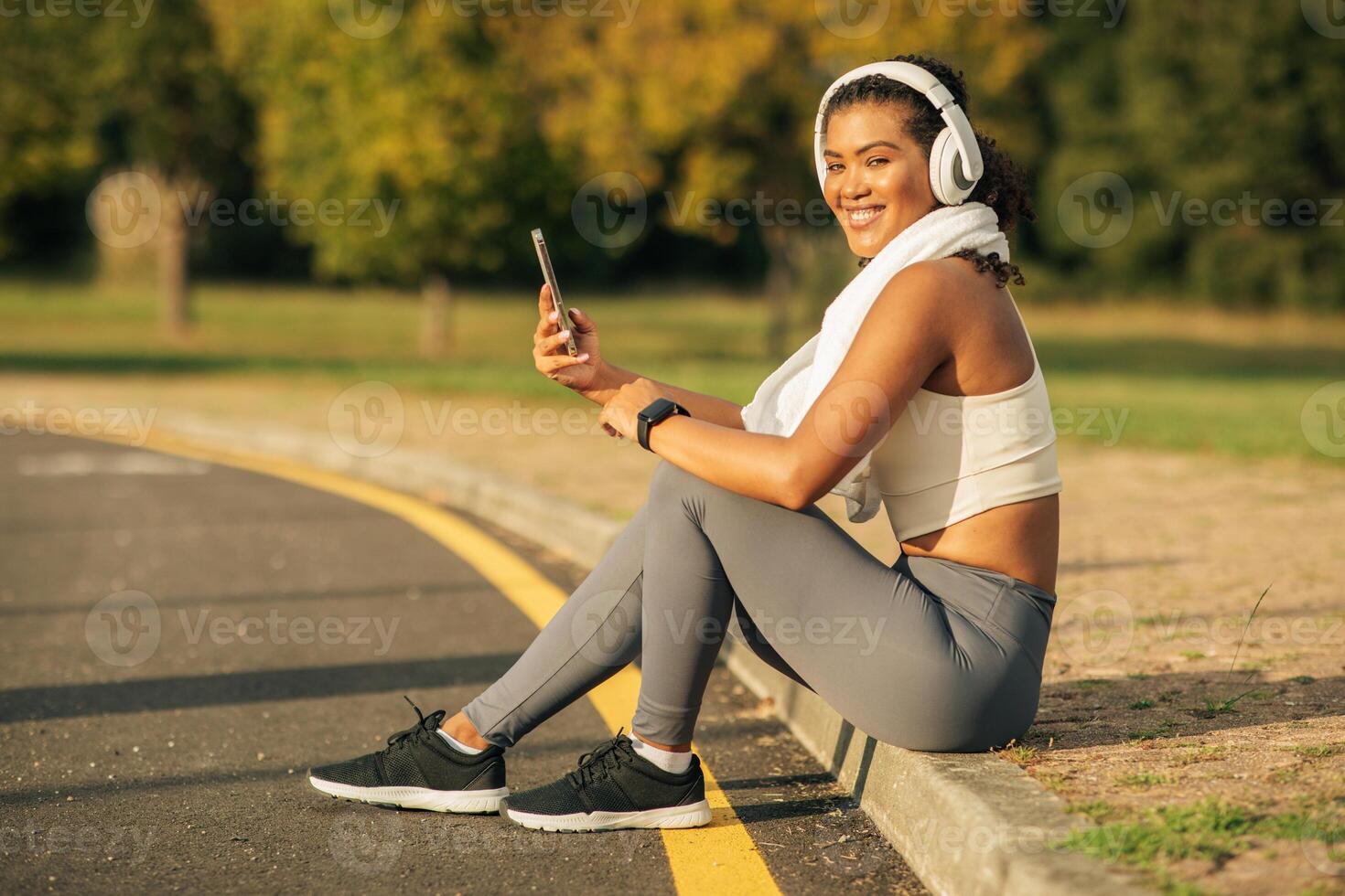 A woman is sitting on the side of a road, holding a cell phone in her hand. She appears to be checking messages or making a call. The background shows a typical road environment. photo
