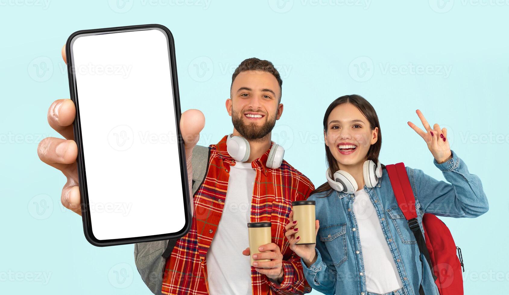 This image features two young, happy students, a man and a woman, holding coffee cups and a smartphone with white screen. They are standing against a blue background and smiling for the camera photo