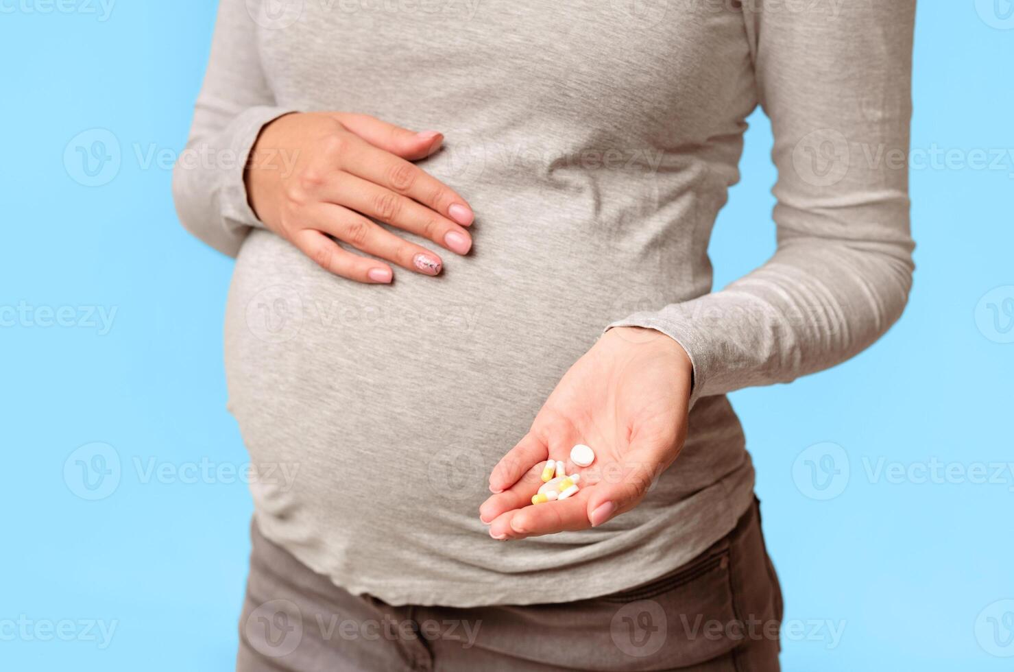 Unrecognizable Pregnant Girl Showing Pills In Hand Touching Belly Standing Over Gray Studio Background. Cropped photo