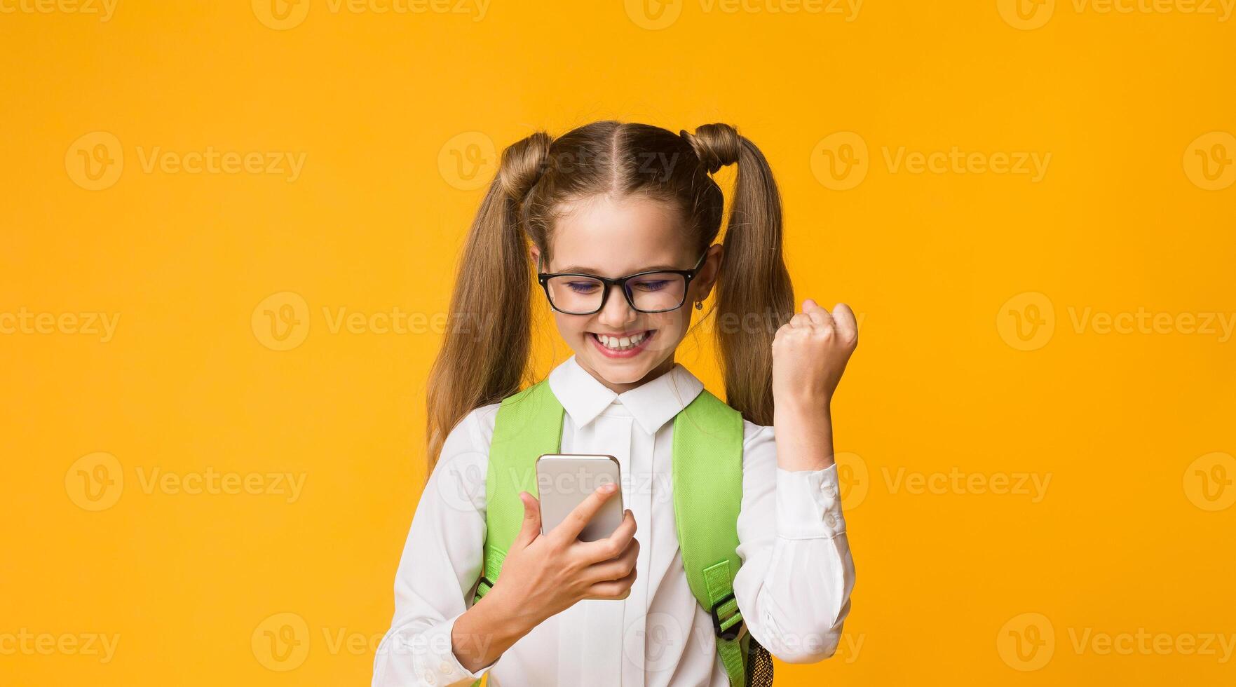 School App. Joyful Schoolgirl Using Mobile Phone Gesturing Yes Passing Test Online Over Yellow Studio Background. photo