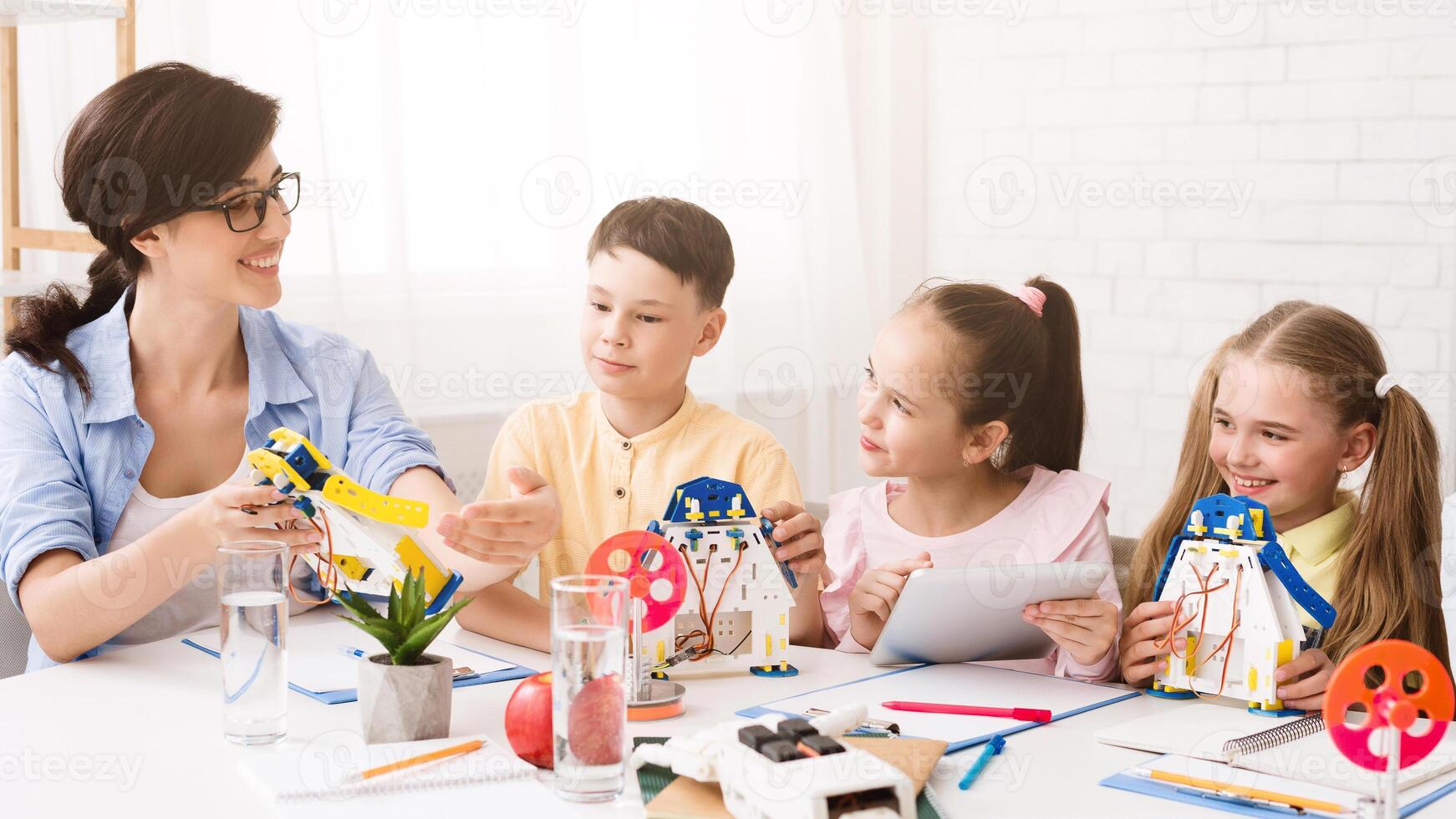 A teacher is showing three students how to build a model robot in a classroom setting. The students are all engaged and interested in the activity. The teacher is pointing to a part of the robot photo