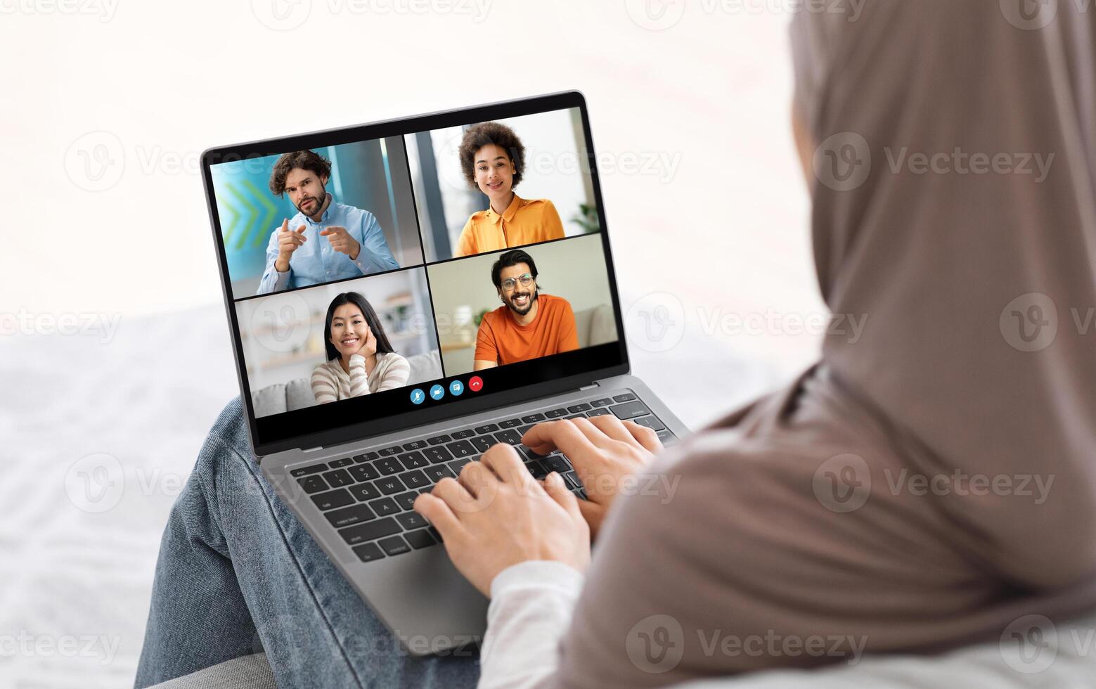 Muslim woman sits on a bed with her back to camera, using laptop to chat with three friends. She is wearing hijab and is typing on keyboard, actively participating in online conversation. photo