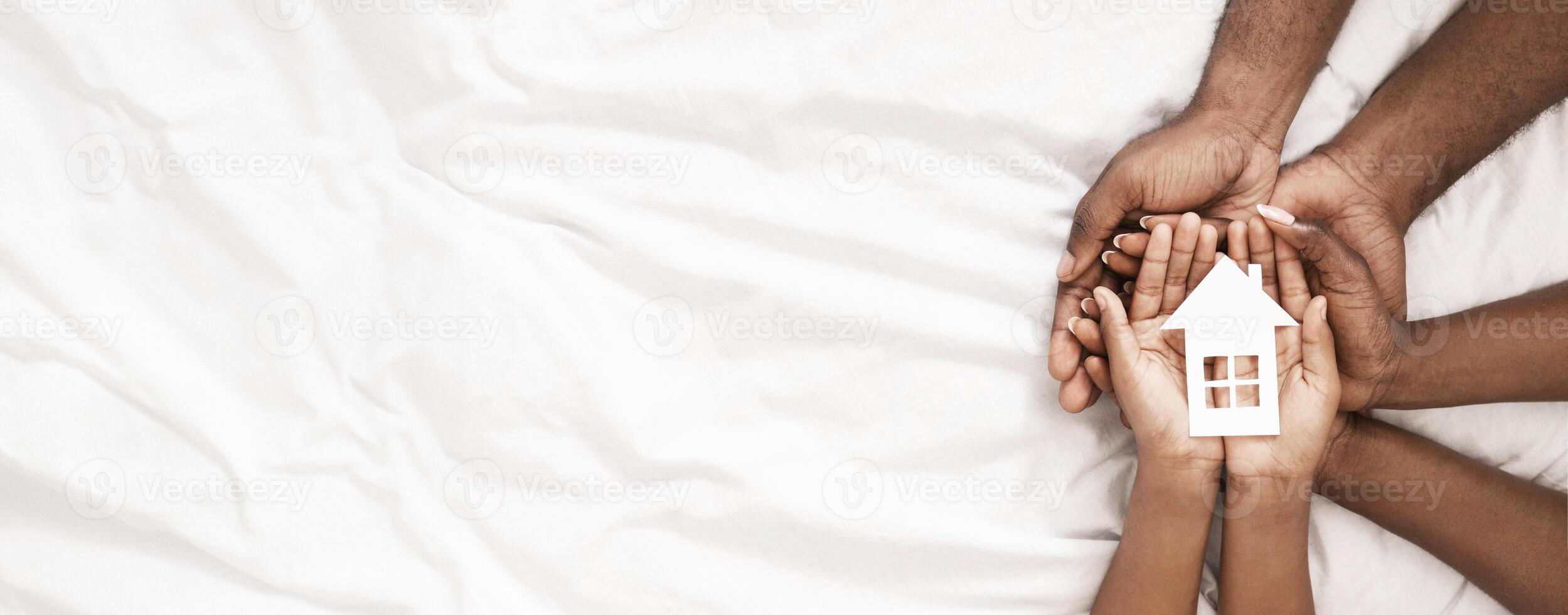 Adoption and family housing concept. Cutout paper house in hands of african american family over white bed sheet background. Closeup, panorama photo