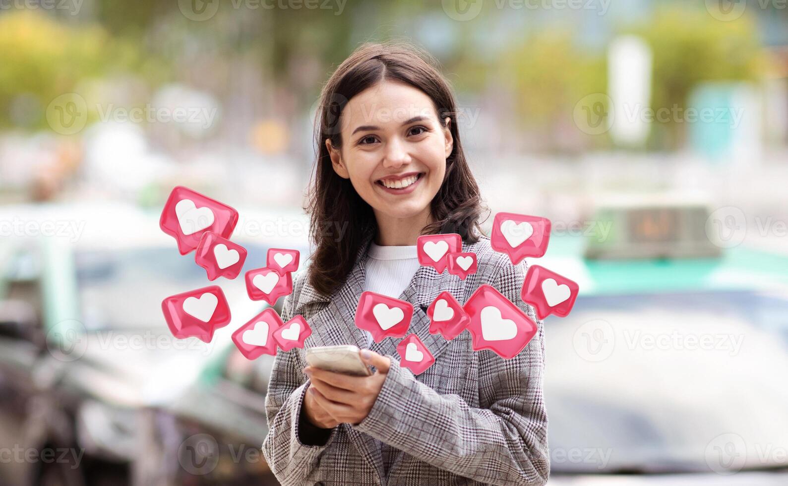 A young woman with brown hair, wearing a gray jacket, smiles and looks directly at the camera as she uses her smartphone. Around her are multiple heart icons in various sizes and positions photo
