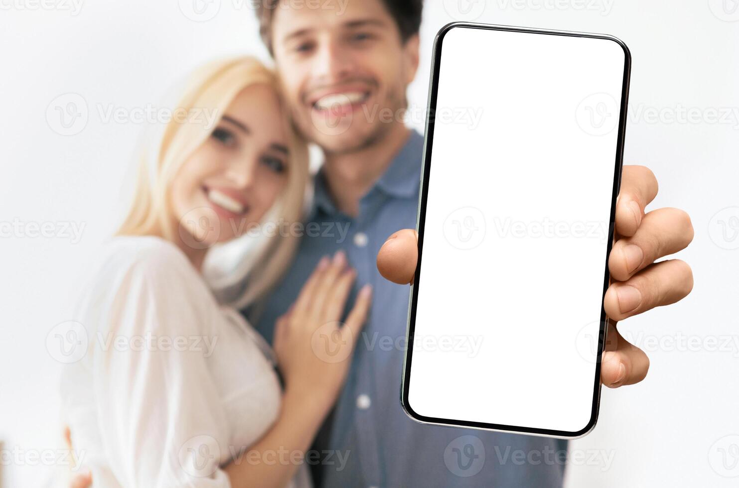 A couple is smiling and looking at the camera as a hand holds a smartphone with a blank white screen in the foreground. photo