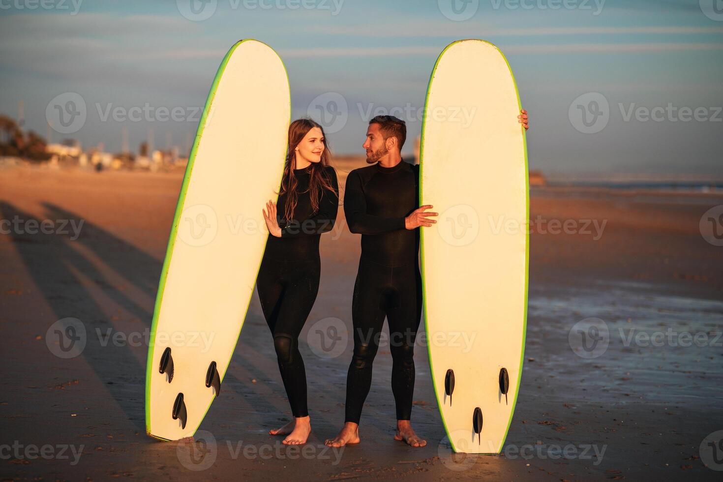 A young couple stands on a sandy beach at sunset, holding their surfboards. The sun is setting in the background, and the sky is a beautiful orange and pink. photo