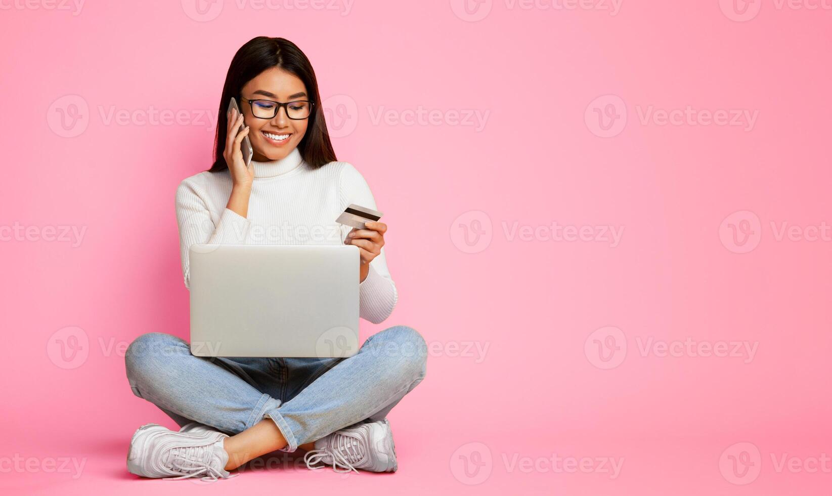 Asian woman sits cross-legged on a pink background. She is holding a credit card in her left hand and talking on a phone with her right hand. She is looking down at a laptop in her lap. photo