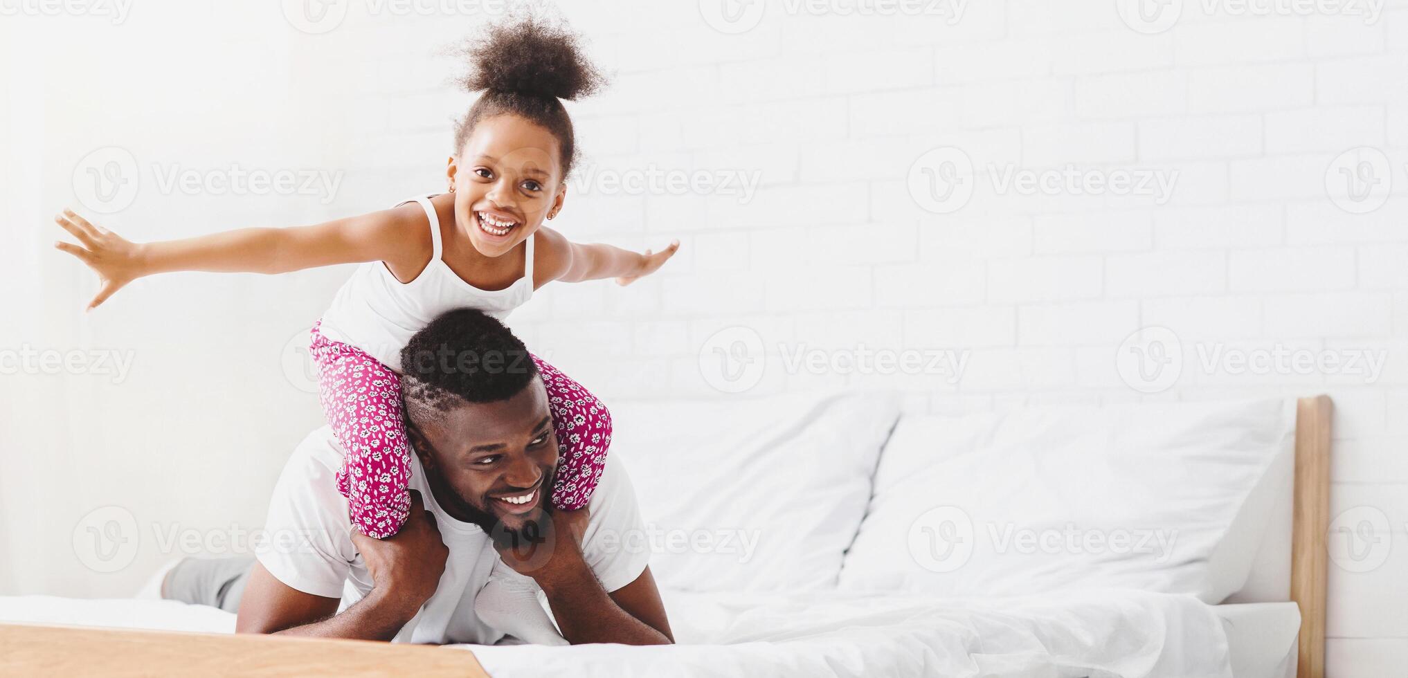 A little black girl with curly hair is riding on her fathers shoulders. Both are smiling and looking at the camera. They are on a white bed with white sheets and pillows, copy space photo