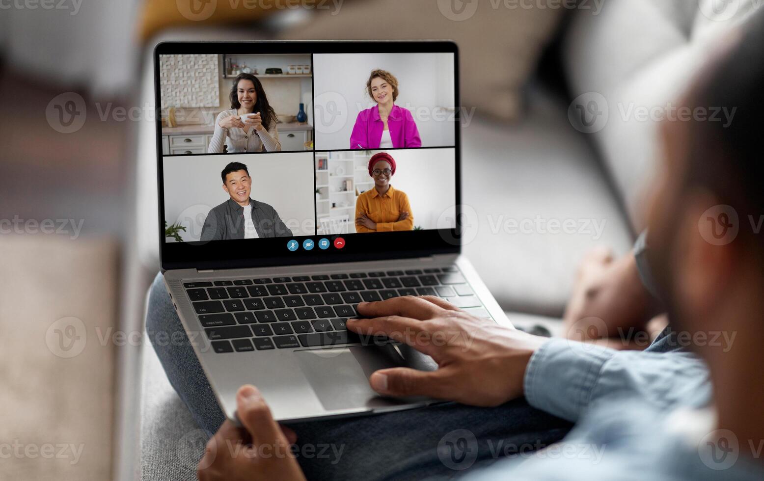A man sits on a couch with a laptop in his lap. The screen displays a conference, showing four individuals in separate squares. The man is using his right hand to interact with the laptop photo