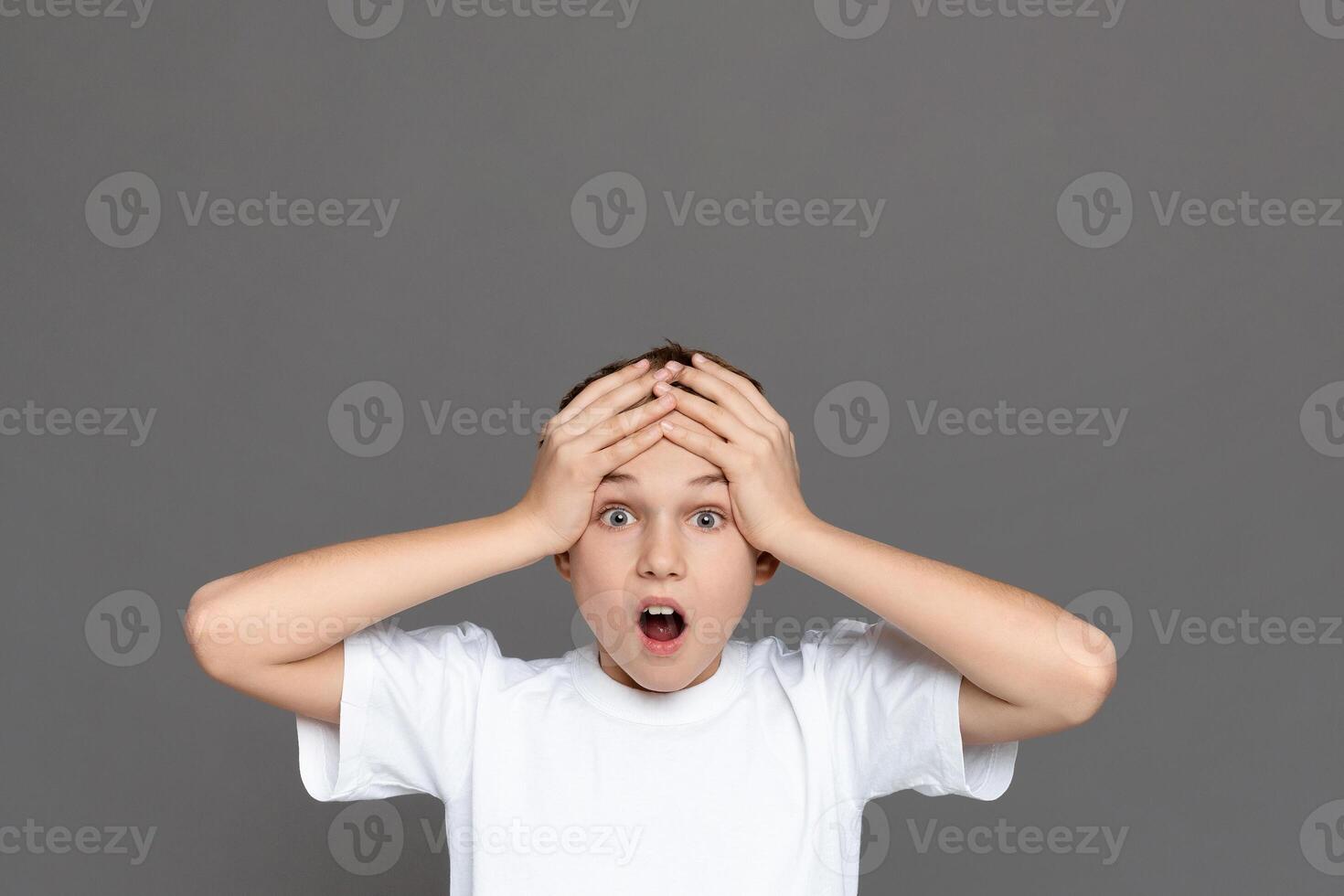 Teenage boy with an expression of sheer surprise, hands on head, isolated on a grey background, depicting a youngster's astonishment photo