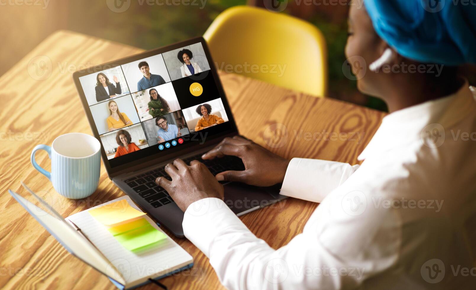 Black woman uses her laptop to engage in a virtual business meeting with multiple colleagues. She sits at a wooden desk with a notebook, pen, and mug beside her at coffee shop photo
