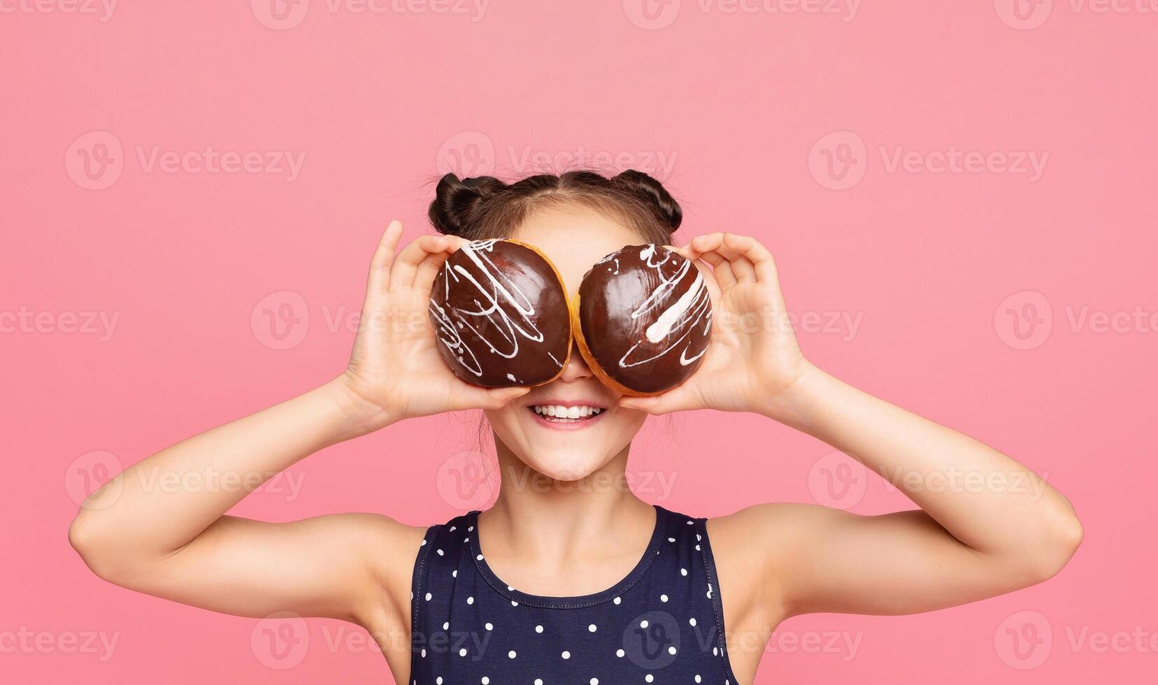 A cheerful young girl with a playful expression holds two chocolate donuts over her eyes against a vibrant pink background. She is wearing a polka dot dress. photo