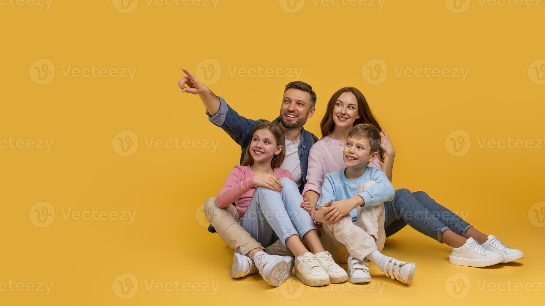 A joyful family is sitting closely with a father pointing at something out of frame, creating a moment of togetherness and curiosity, pointing at copy space on yellow background photo