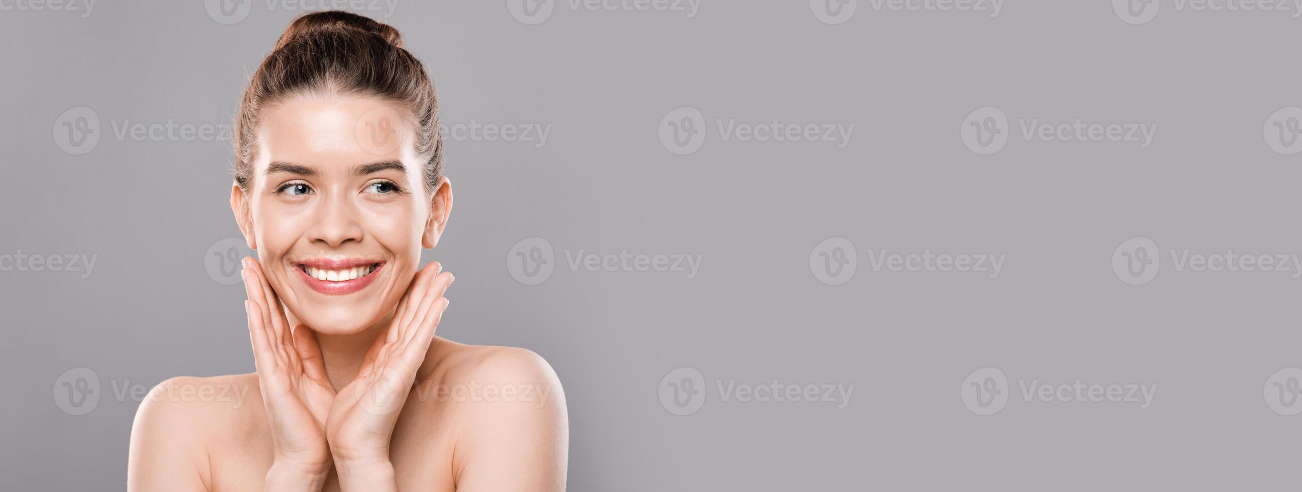 Face care concept. Cheerful young woman touching her face and looking at copy space over gray studio background photo