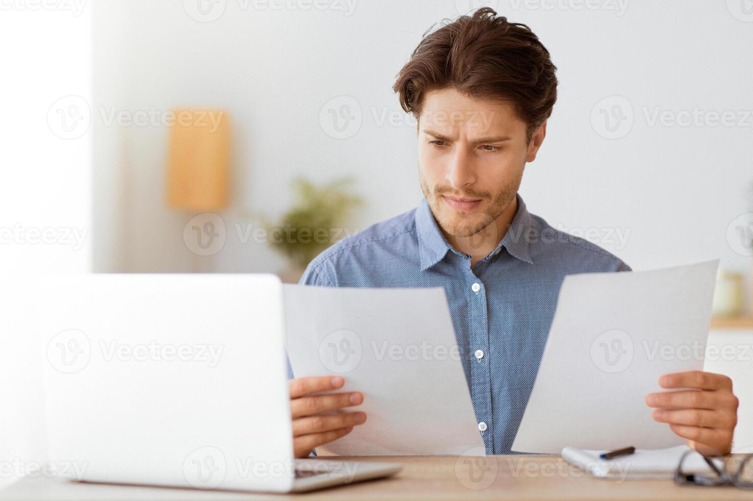 A man in a blue shirt reviews paperwork while sitting at his desk with a laptop and other office supplies. photo
