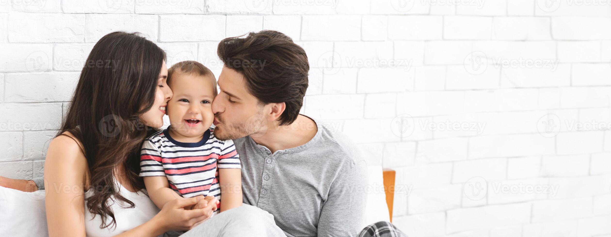 A joyful family is enjoying a tender moment in their cozy living room. The parents are sharing affectionate kisses with their smiling baby, who looks delighted amidst the warmth of love and comfort photo
