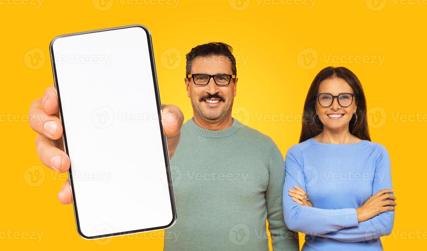 Mature man and woman are standing in front of a yellow background. The man is holding a smartphone with a blank white screen towards the camera photo