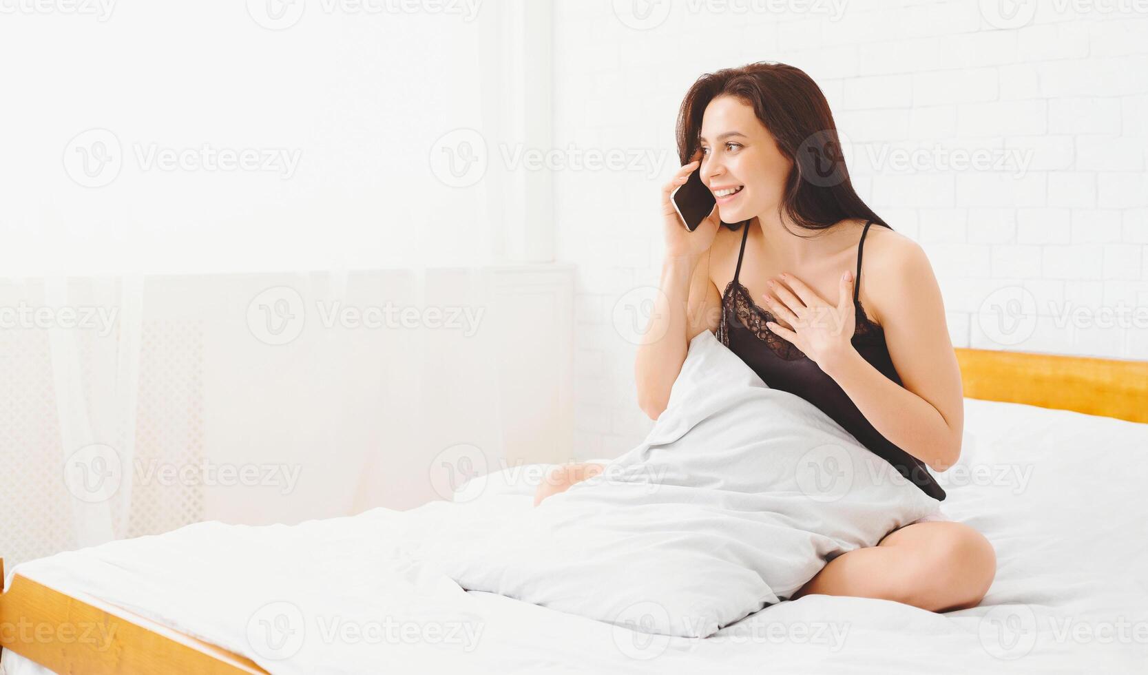 A woman is sitting on a bed, holding a cell phone to her ear and engaged in conversation. She appears focused and leaning slightly forward, with a neutral expression on her face. photo