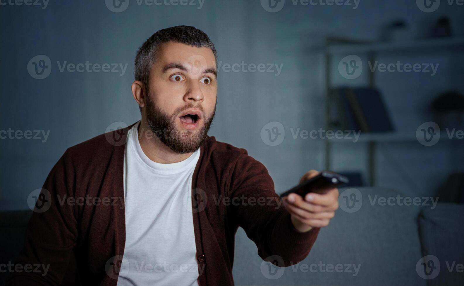 A man with a shocked expression is sitting on his couch at home in the evening, holding a TV remote and watching an unexpected scene on the screen. photo