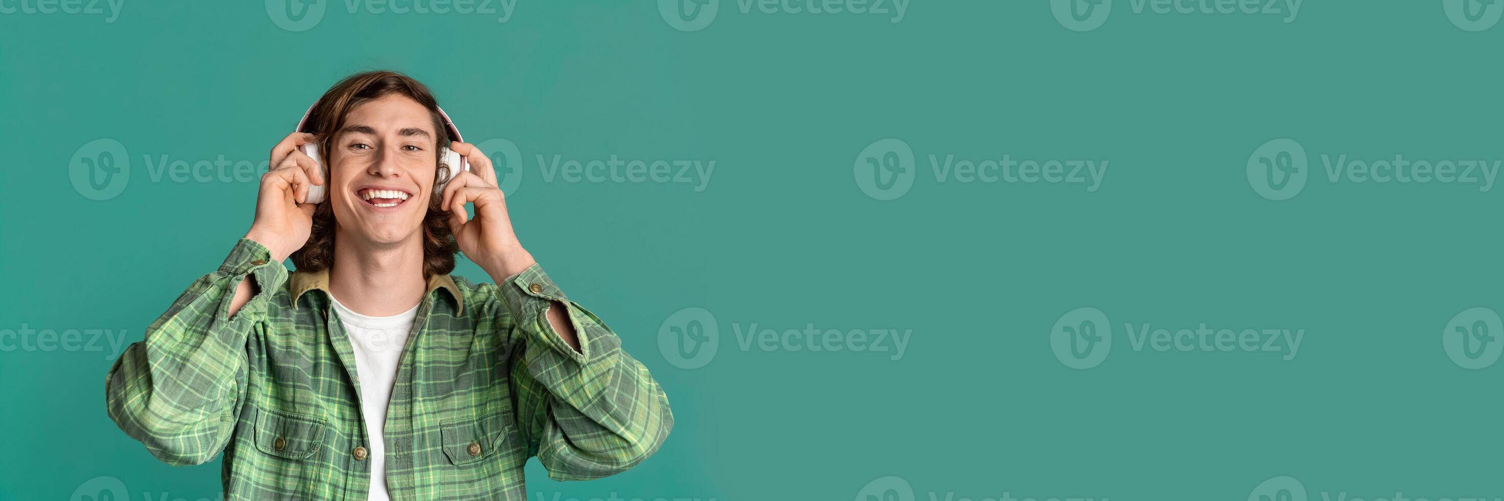 Music everywhere. Cheerful young guy with headset listening to favorite song on turquoise studio background, panorama with copy space photo