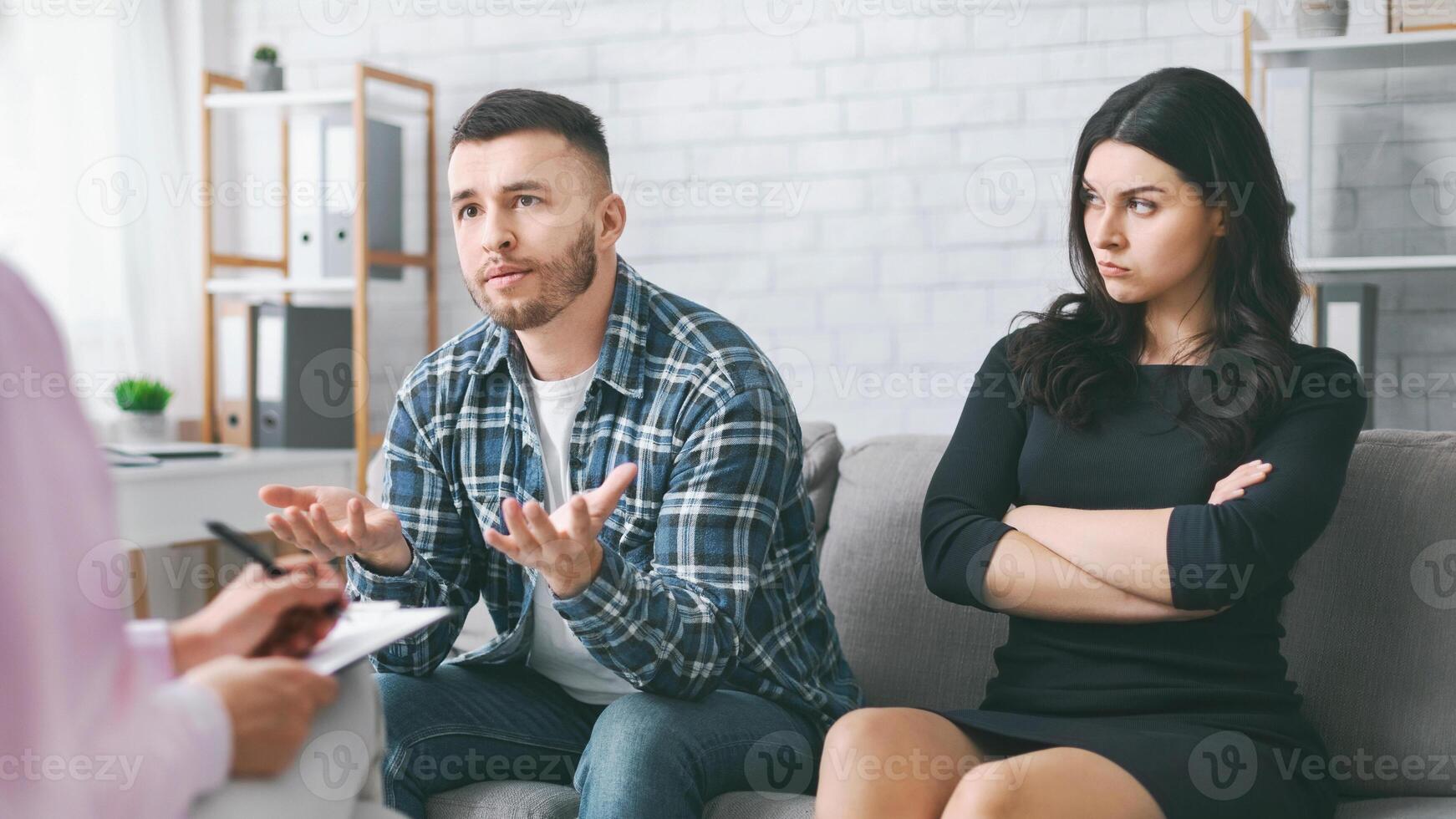 A couple sits on a couch in a counseling session. The man, wearing a plaid shirt and jeans, is speaking with animated gestures photo