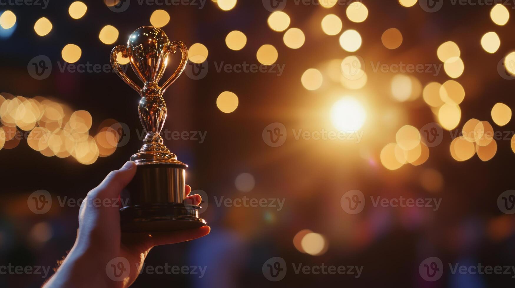 A hand proudly holds a shiny trophy, illuminated by a backdrop of soft, glowing lights during a festive celebration. photo
