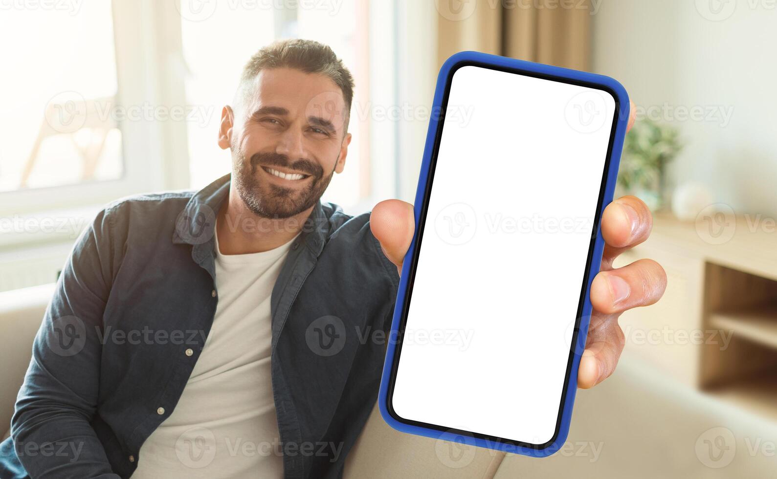 A man is seated on a couch while actively holding up a cell phone in his hand. He appears focused on the devices screen, engrossed in whatever content he is viewing. photo