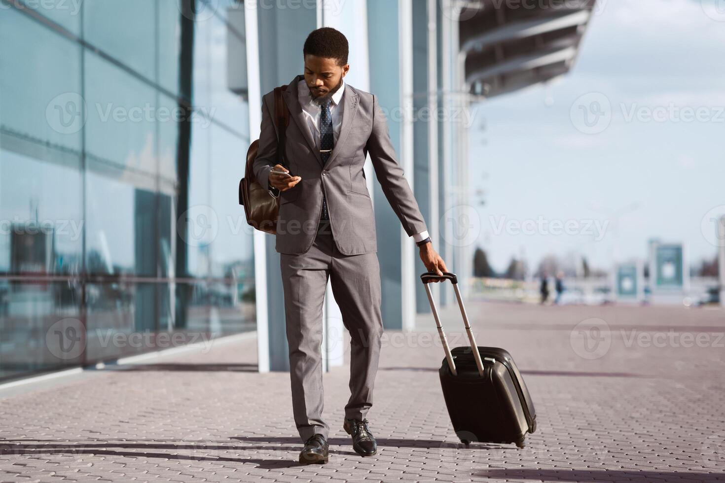 Black businessman is using his phone while standing with luggage outside a modern airport. The mood is busy and professional, capturing a moment of travel and communication. photo
