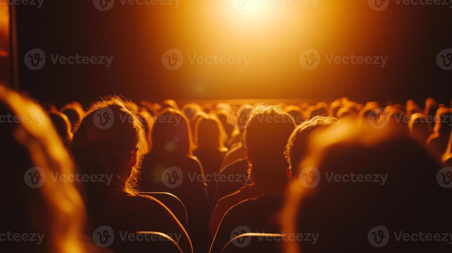 A large crowd is seated in a theater, facing a brightly lit stage that illuminates the darkened room. photo