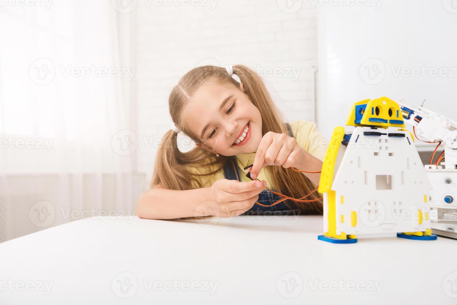 A young girl with pigtails smiles as she focuses on connecting wires to her robot. The robot is white and yellow, and the girl is sitting at a table in a brightly lit room. photo
