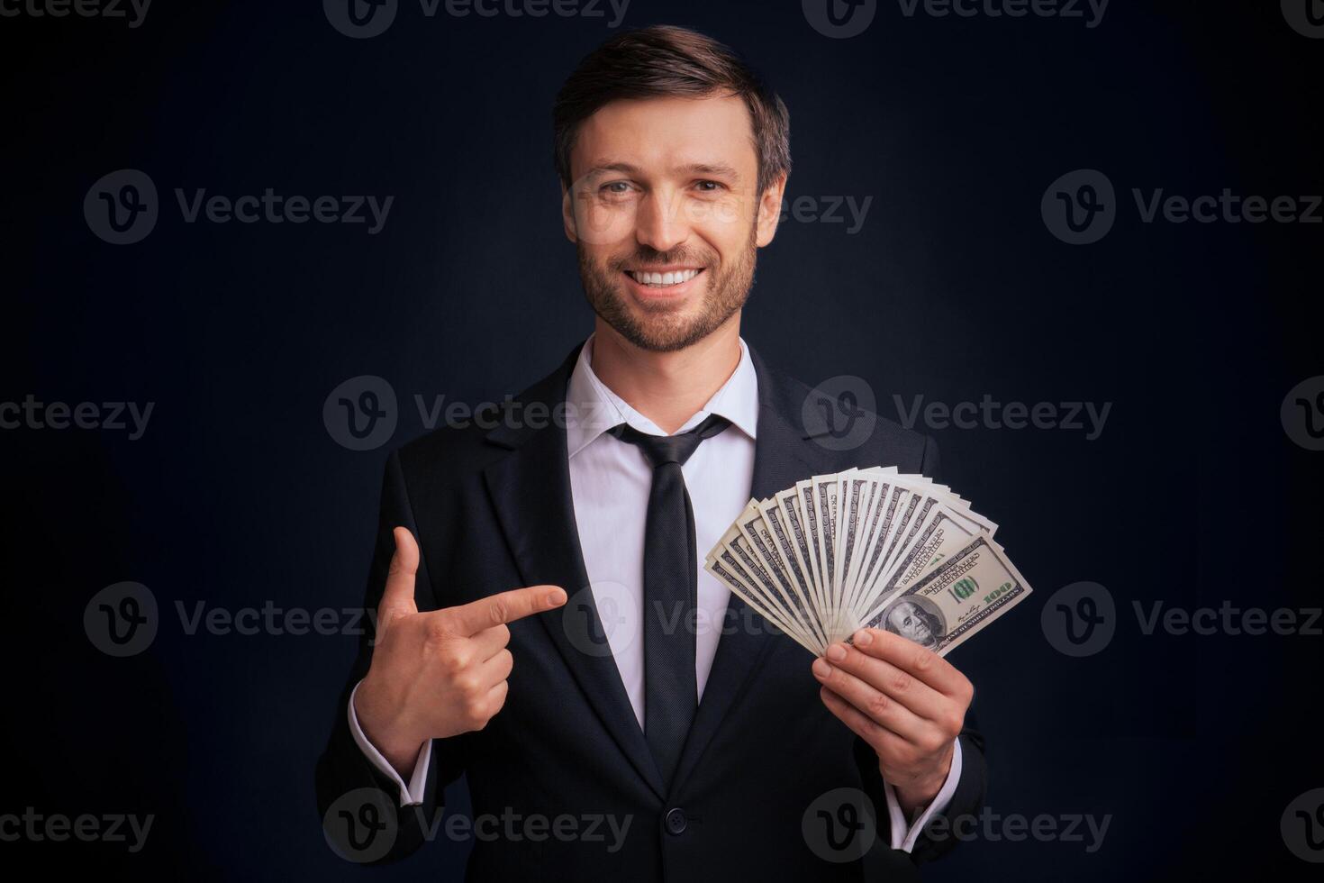 Income. Smiling Businessman Pointing Finger At Money Cash In His Hand Standing Over Black Studio Background. photo