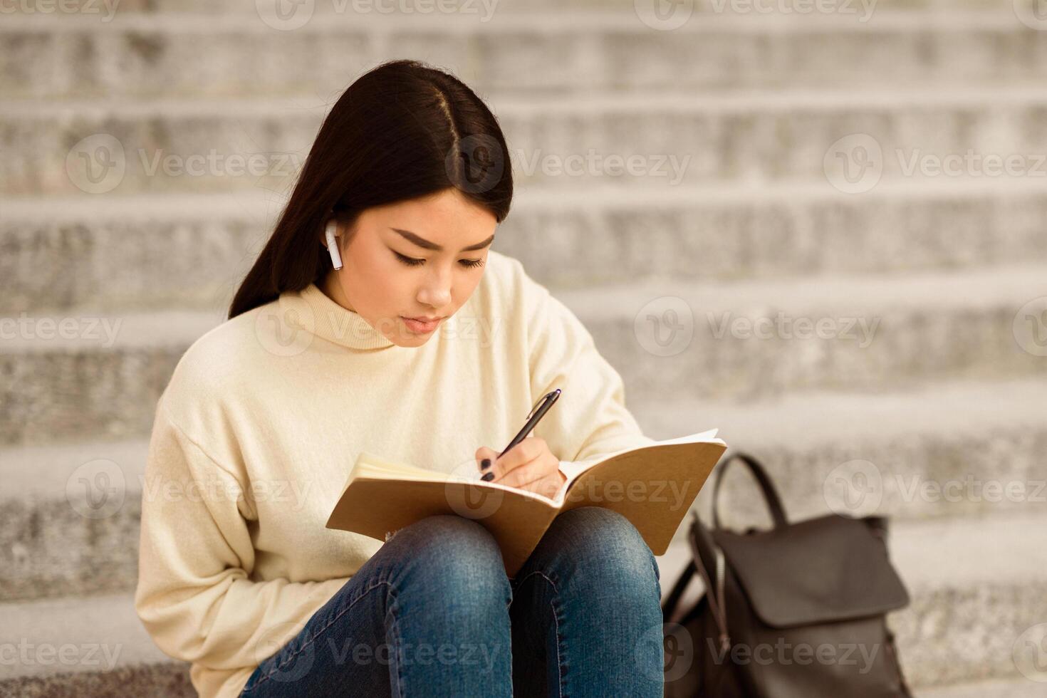 Chinese woman is seated on a flight of steps, engrossed in writing in a notebook. Her posture is attentive, pen in hand, as she fills the pages with her thoughts. photo