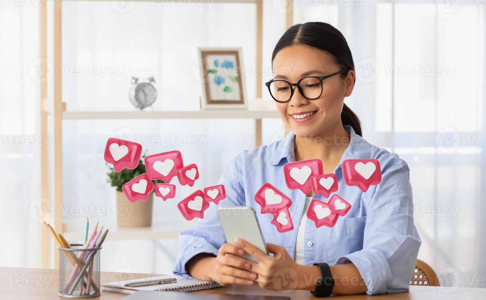 A smiling Asian woman sits at her desk, looking down at her phone while holding it with both hands. Pink heart icons float around her, suggesting she is receiving likes and positive engagement photo