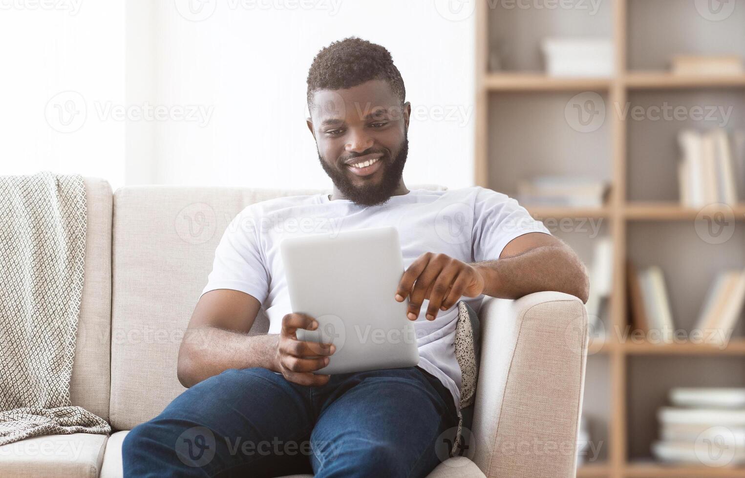 African American man sits on a white couch in a living room, smiling as he uses a tablet. He is wearing a white t-shirt and blue jeans, and the couch is next to a bookshelf. photo
