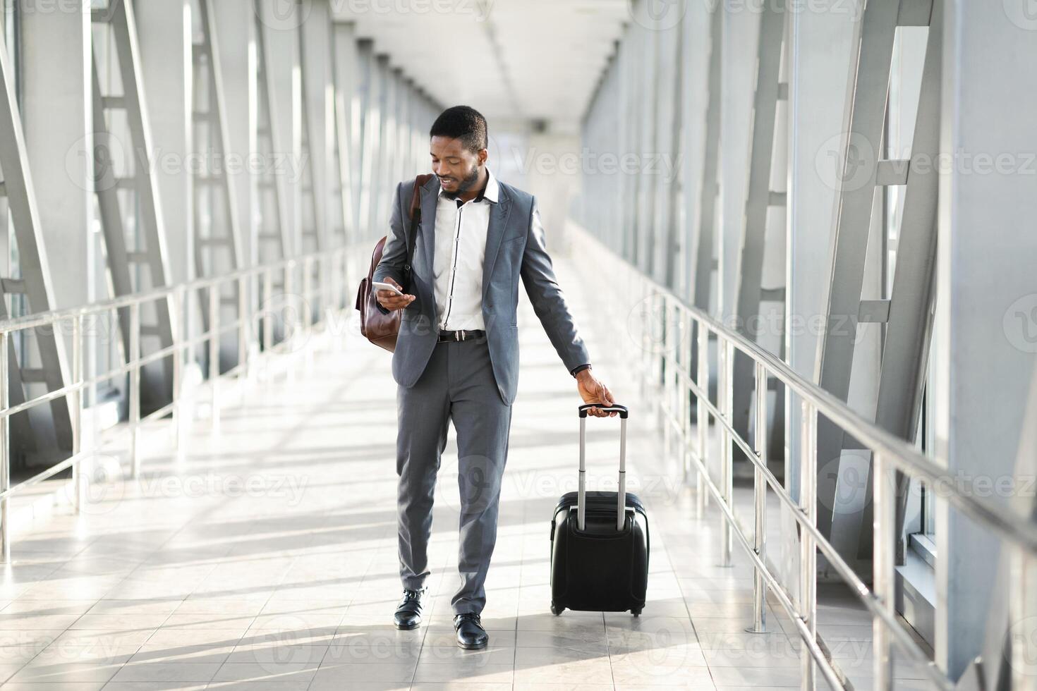 Black businessman in a suit checking his phone while walking by modern airport with a rolling suitcase, copy space photo