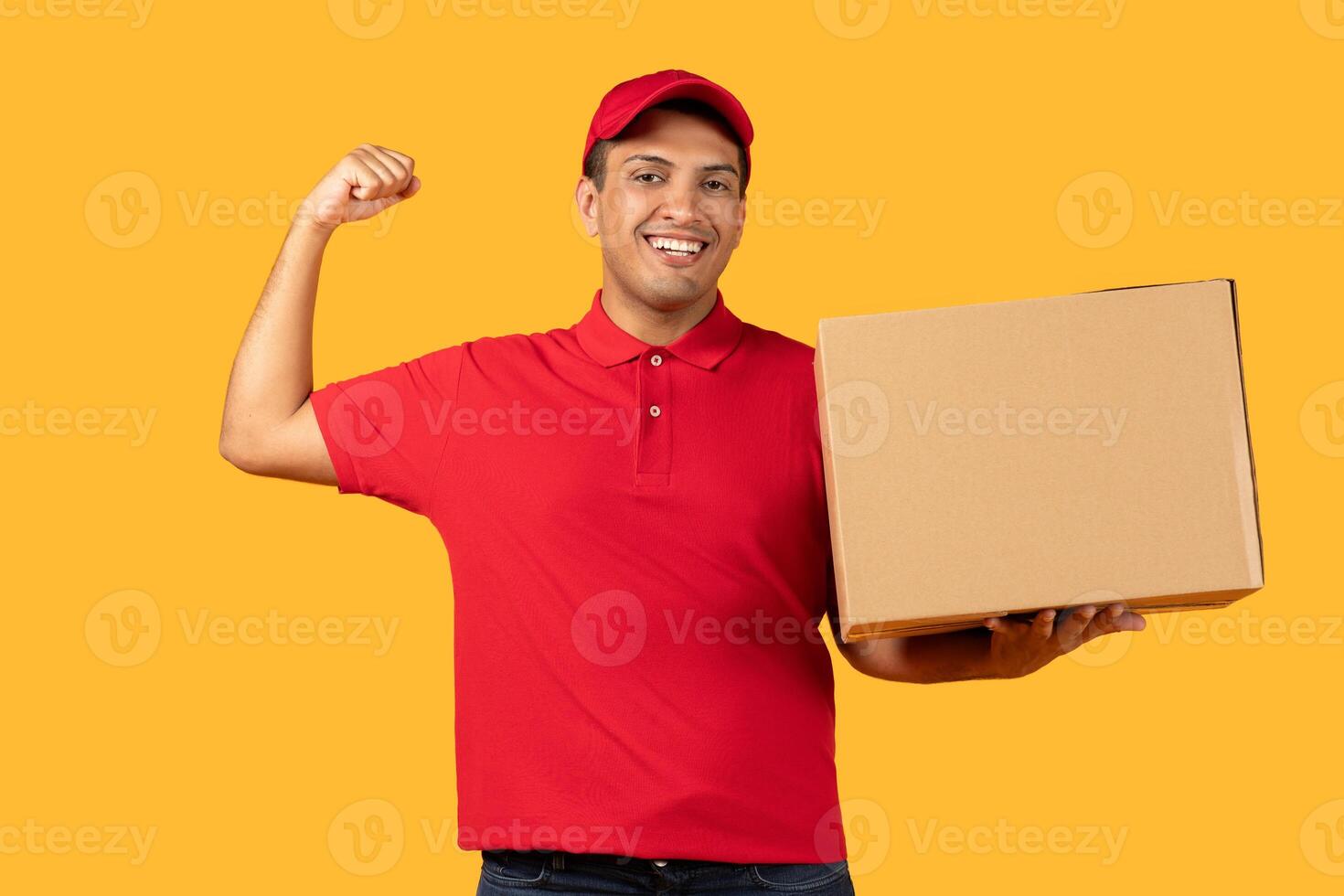 A cheerful delivery worker man in a red cap and polo shirt holds a cardboard box while flexing his arm, showing enthusiasm and strength. photo
