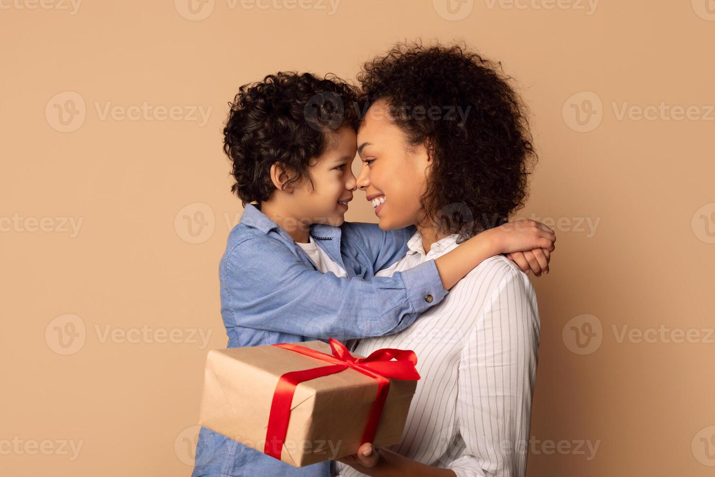 Cute African American little boy greeting his mom with Mothers Day, giving her wrapped box and embracing his mummy, beige studio background photo