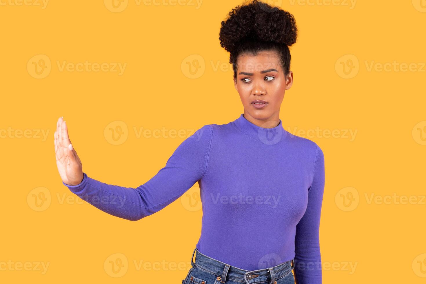 A young woman stands confidently against a bright yellow backdrop. She extends her hand forward, signaling a clear gesture of dismissal. Her expression suggests assertiveness photo