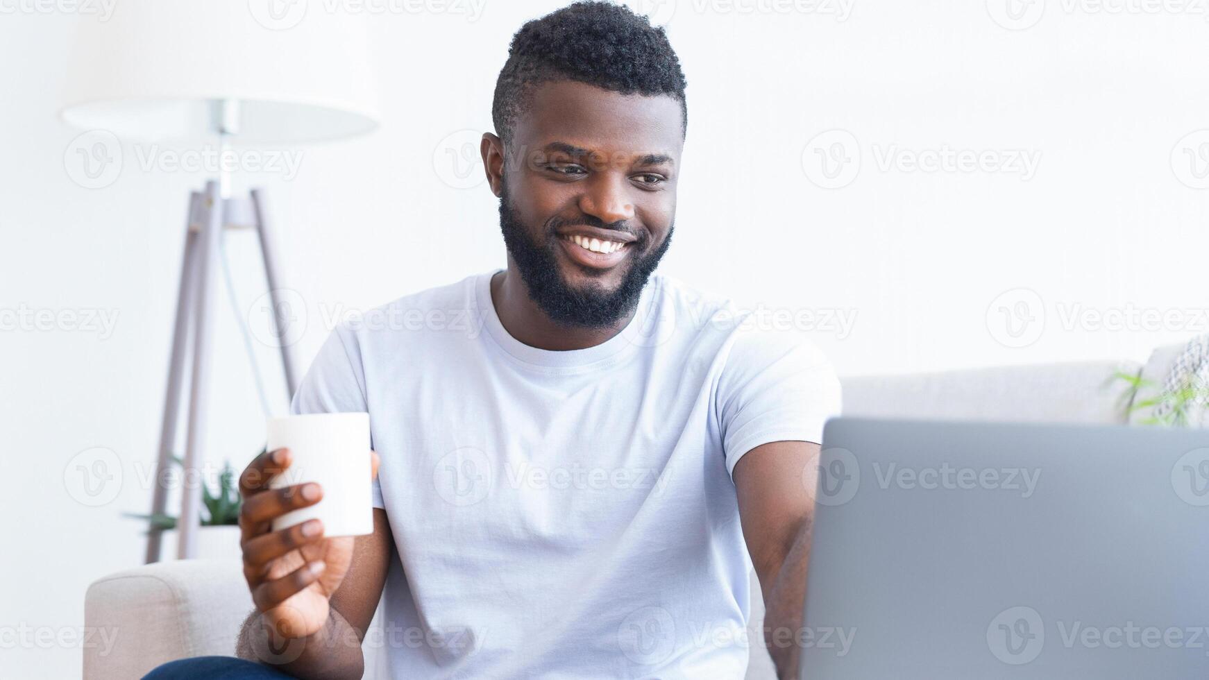 Black man is sitting on a couch in his home, smiling and using a laptop. He is holding a white mug in his right hand. The man is wearing a white t-shirt and appears to be enjoying his work. photo