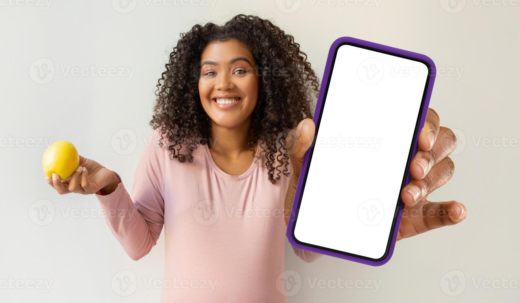 African American woman is holding a cell phone in one hand and an apple in the other hand. She appears to be looking at the screen of the phone, with the apple held casually. photo
