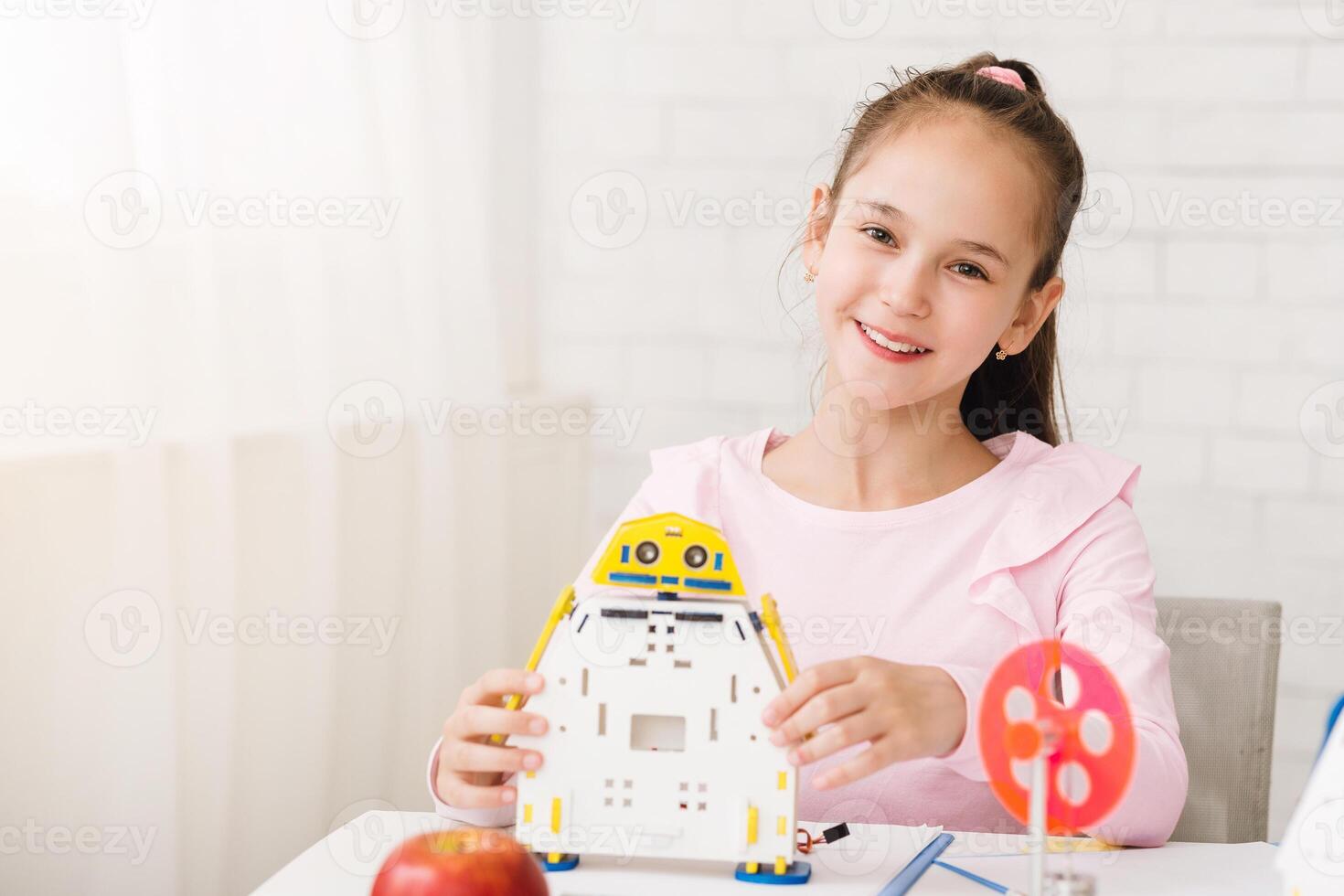 A young girl with a ponytail smiles while holding a robotic toy made of yellow, blue, and white plastic. She is sitting at a table with various other building supplies in front of her. photo
