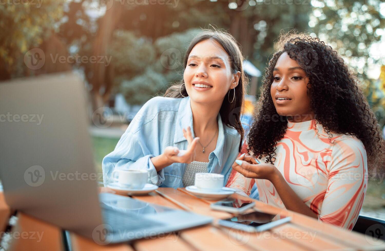 Two women are seated at a table in a cafe setting. One woman is looking at the laptop and speaking, while the other woman listens intently. Both women have coffee cups in front of them. photo