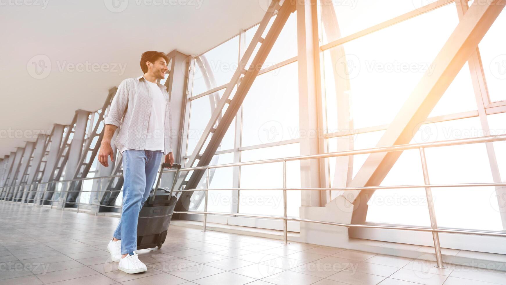 Enjoying vacation. Cheerful man walking with luggage in airport terminal, looking at sunset in window, empty space photo