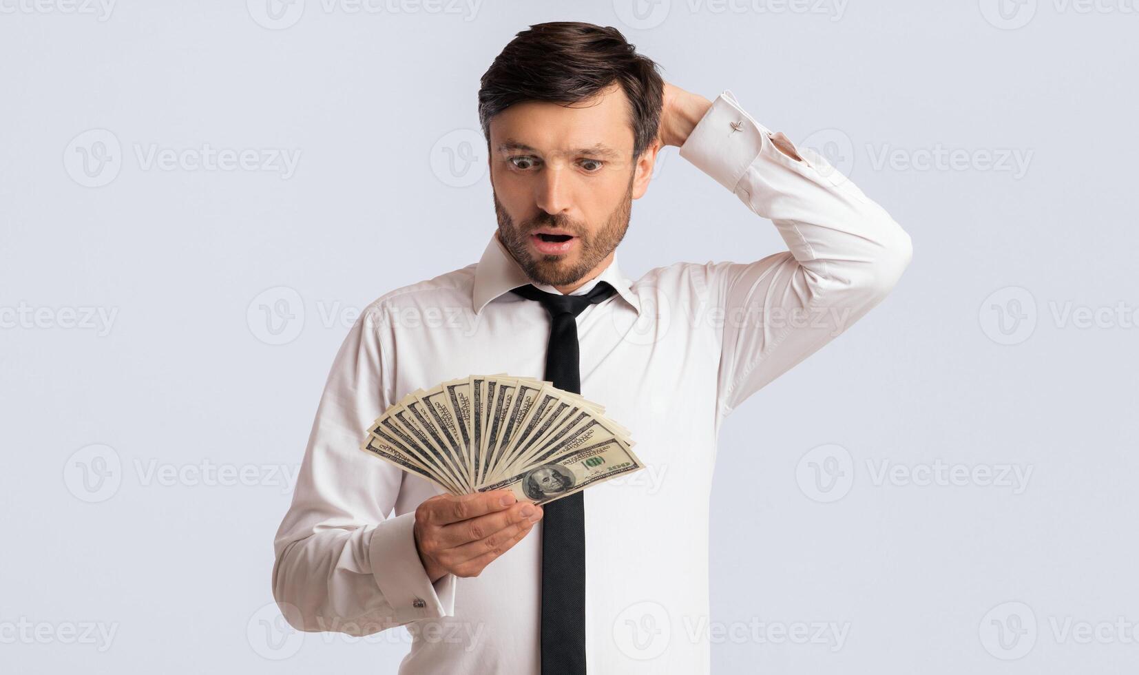 Financial illiteracy. Shocked man holding money scratching his head on white background. Isolated, studio shot photo